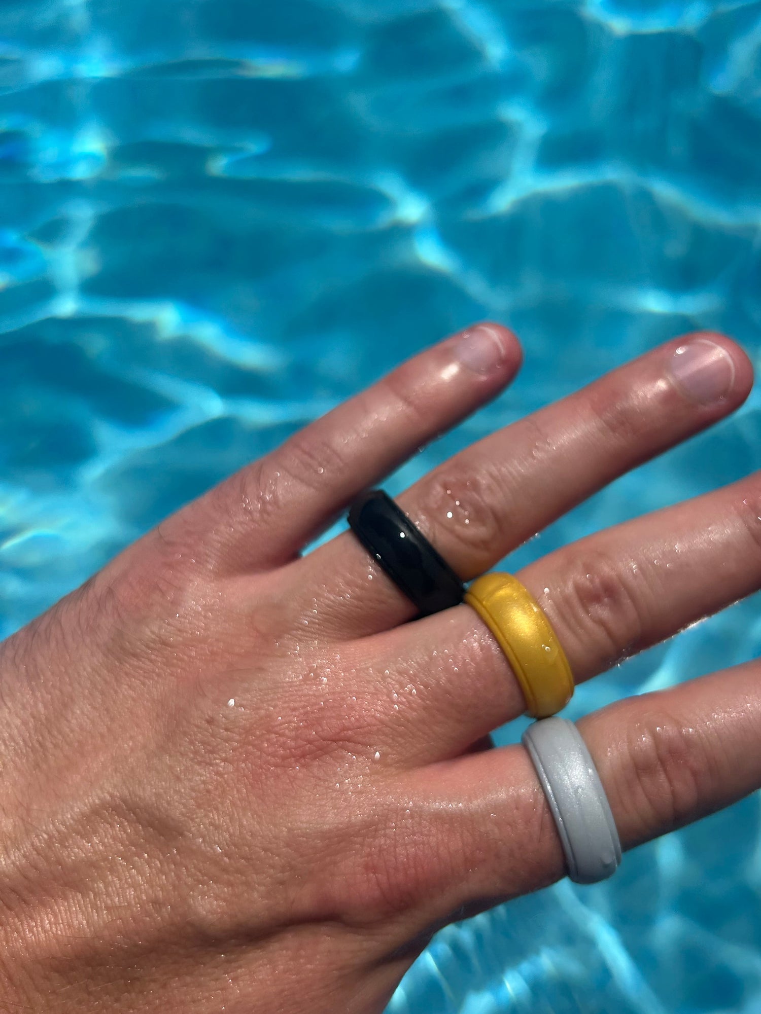 Three minimalist comfort silicone rings in black, yellow, and light grey worn on a person's hand, photographed outdoors over bright blue pool water.